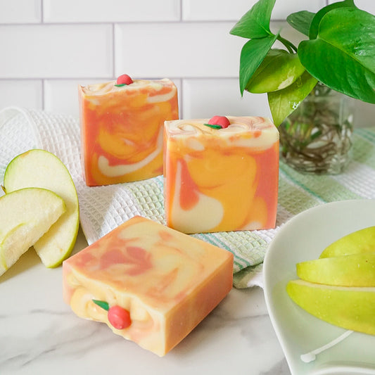 Three square-shaped soap bars with a fruit pattern on a marble surface with a plant in the background.