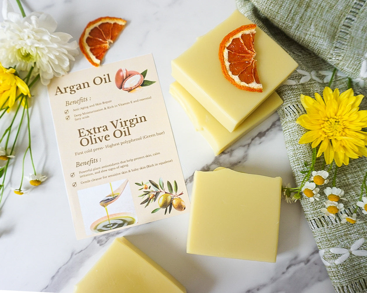 Yellow soap bars with orange slices and a card on a marble surface with flowers and a towel.
