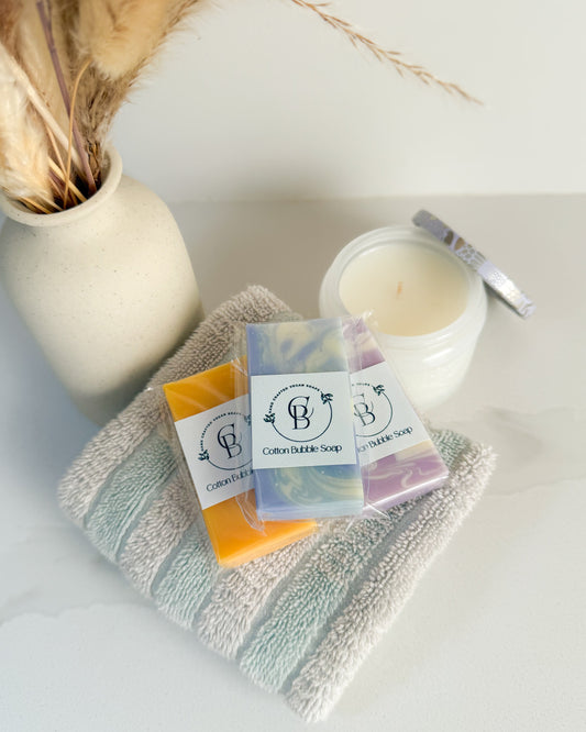 Three bars of soap with a brand label on a towel next to a candle and a vase.