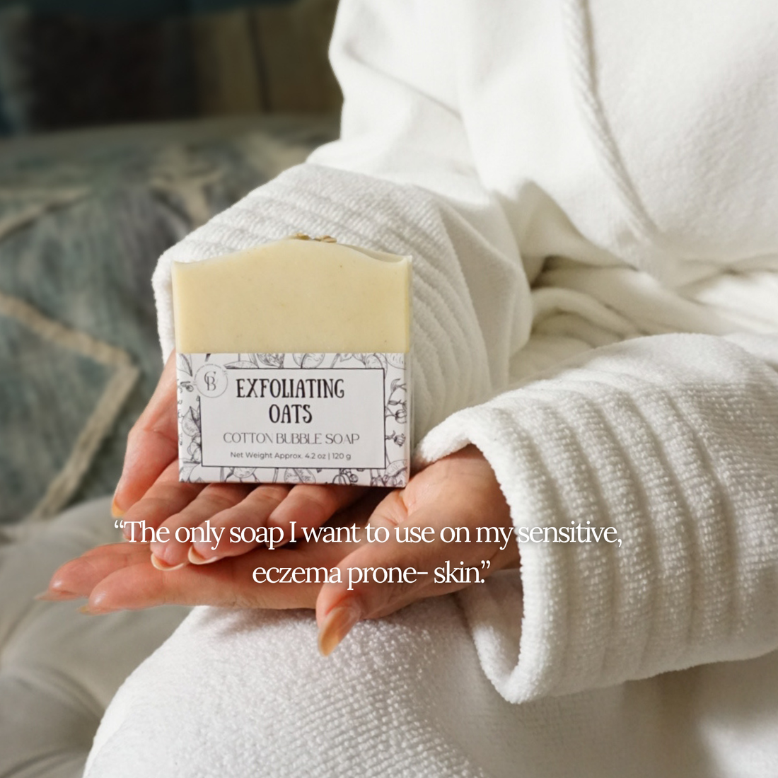 Person in a white bathrobe holding 'exfoliating oats' soap in their hands