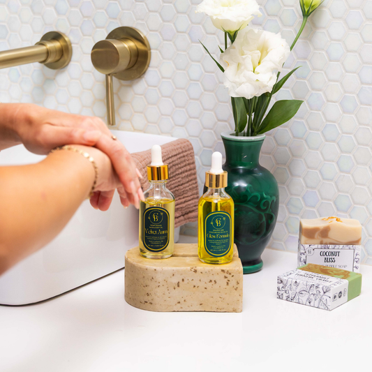 bathroom image with a white flower background in a dark green vase and 2 soaps and 2 glass bottle oils and someone rubbing hands
