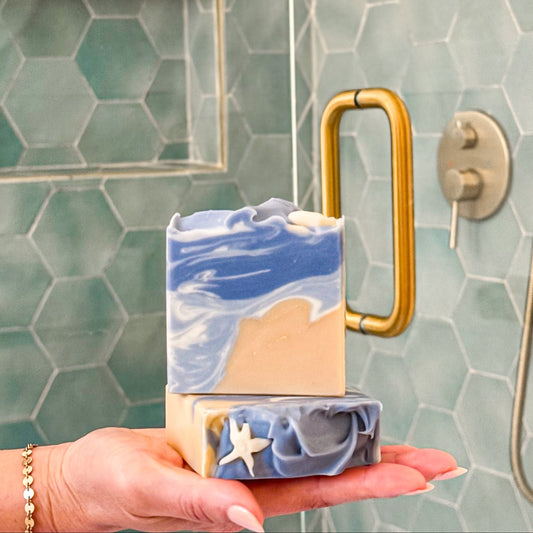 Hand holding a layered soap bar with a gold handle against a tiled shower wall.