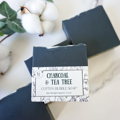 Two bars of charcoal and tea tree soap with a packaging label on a marble surface.
