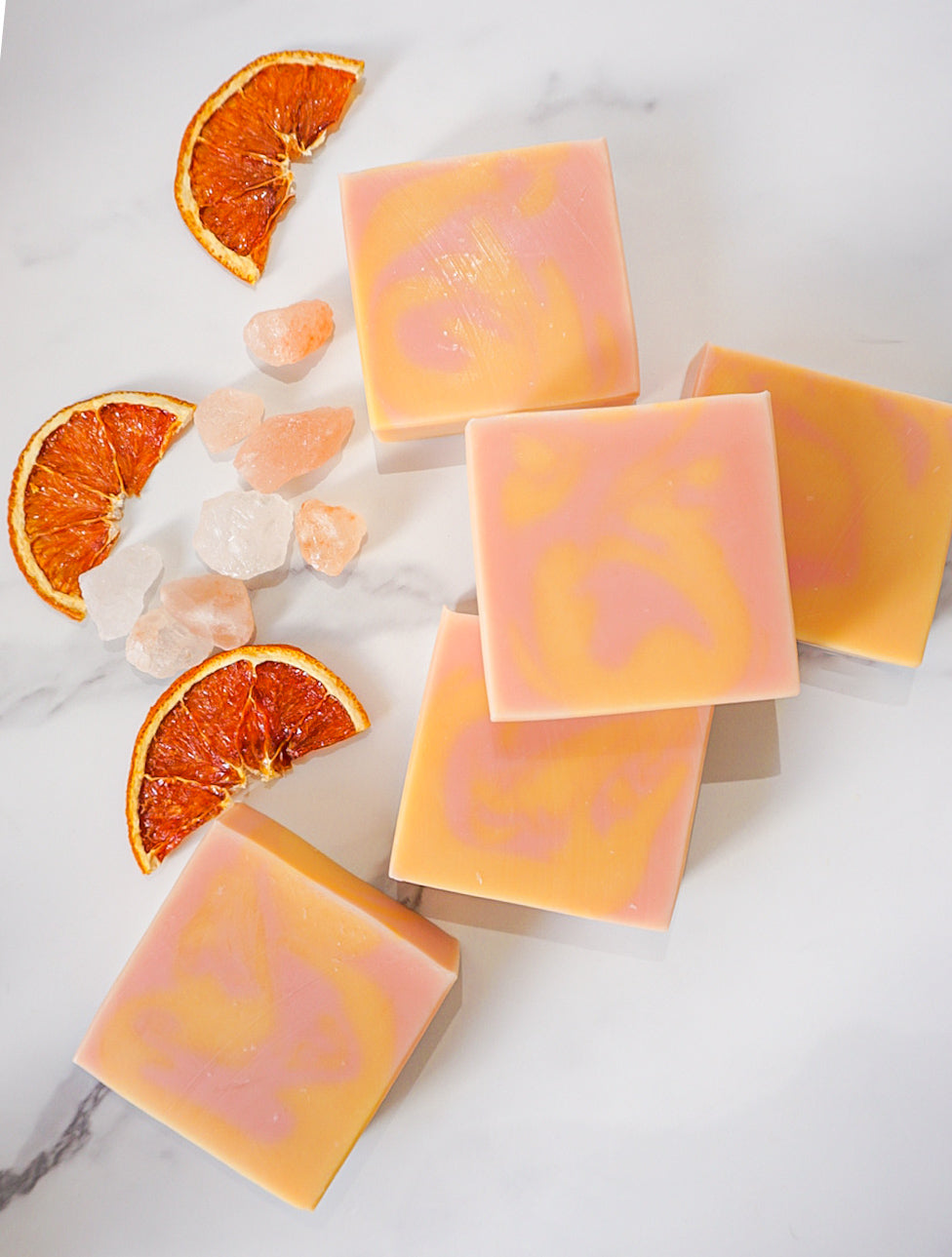 Orange-colored soap bars with orange slices and ice cubes on a marble surface