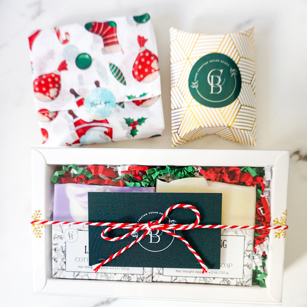 Gift box with festive wrapping paper, cards, and a decorative pillow on a white surface.