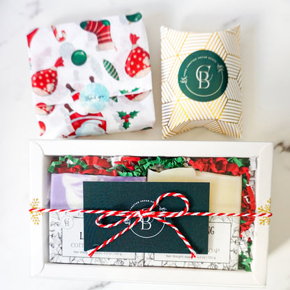 Gift box with festive wrapping paper, cards, and a decorative pillow on a white surface.