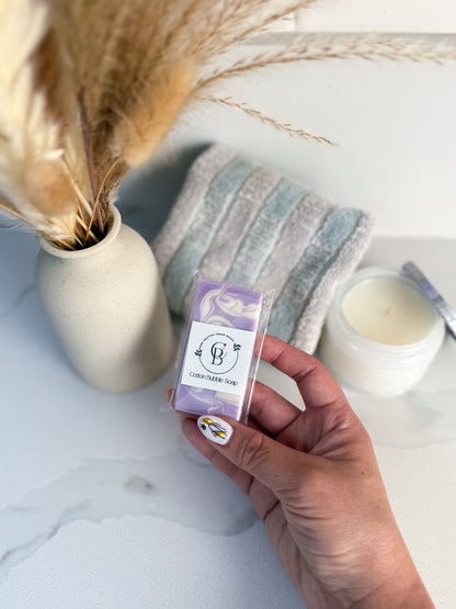 Hand holding a purple soap package with a vase, candle, and towel in the background.