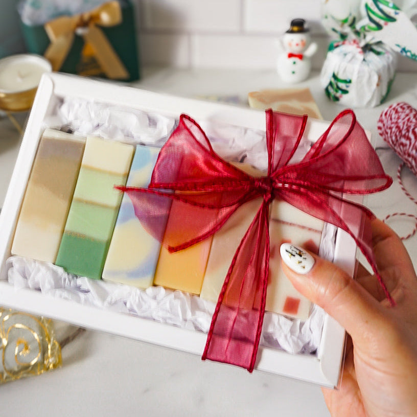 Box of colorful soap bars with a red ribbon held by a hand, with decorative items in the background.