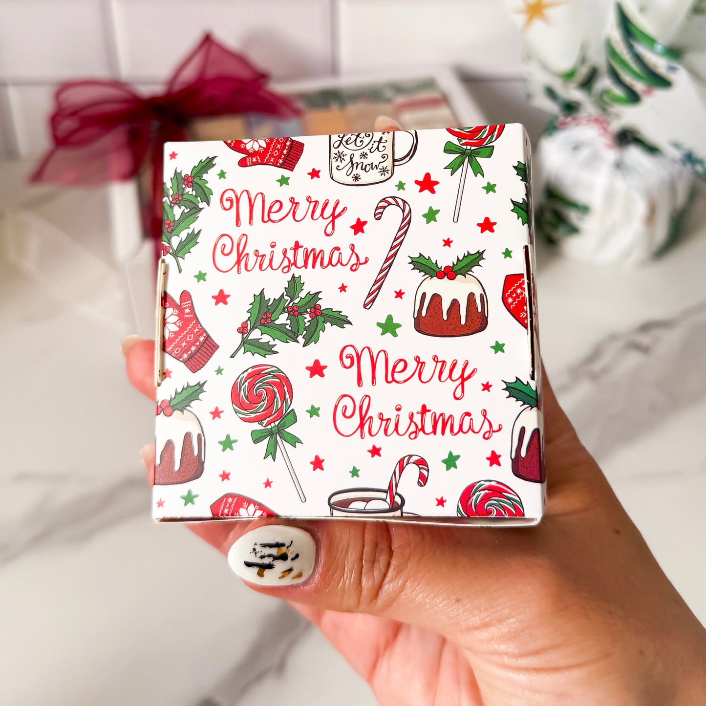 Hand holding a small box with Christmas-themed design featuring candy canes, cakes, and 'Merry Christmas' text.