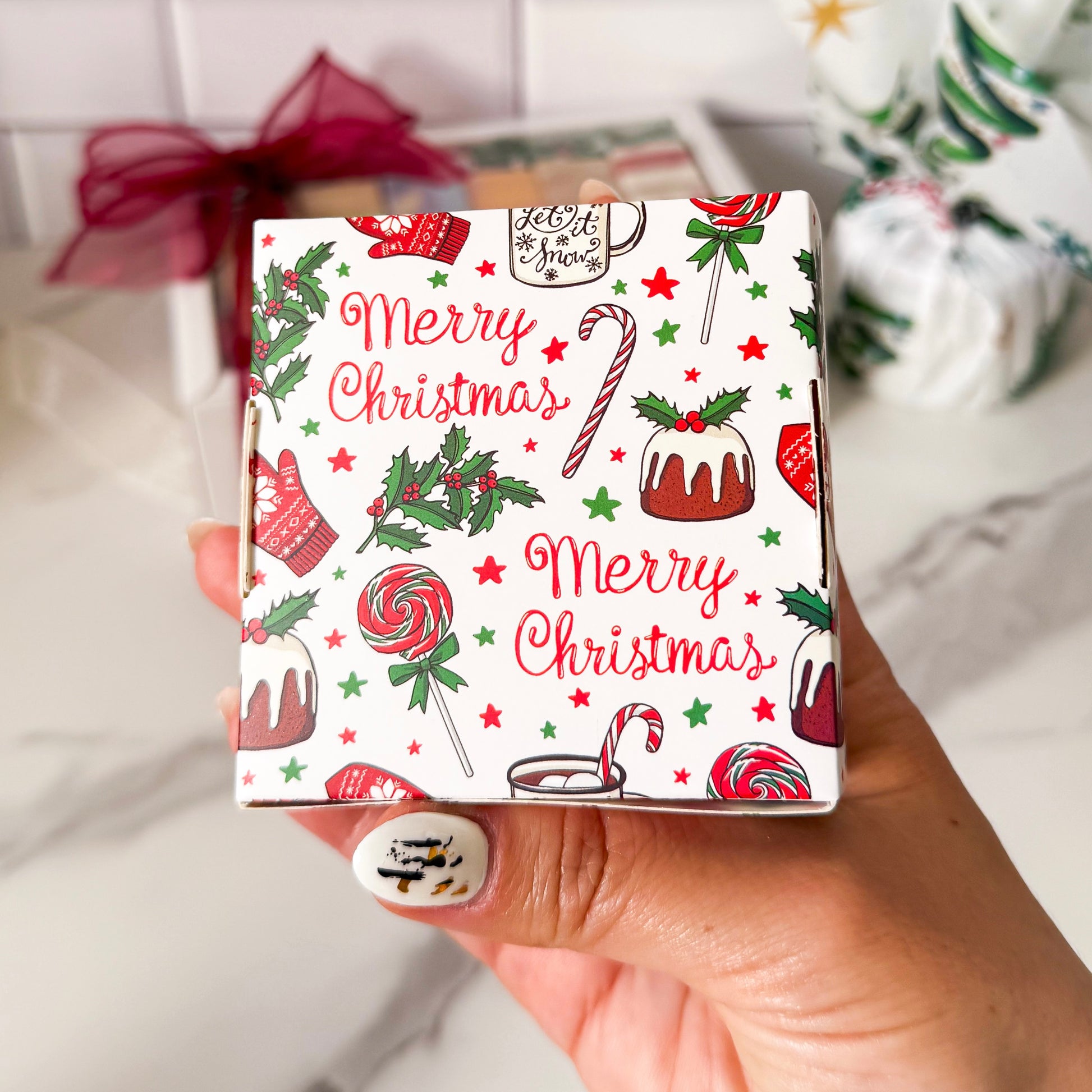 Hand holding a small box with Christmas-themed design featuring candy canes, cakes, and 'Merry Christmas' text.