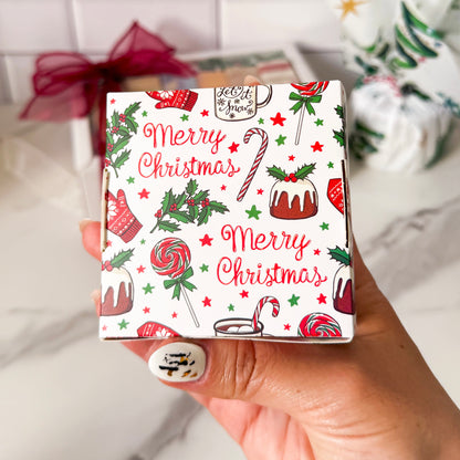Hand holding a small box with Christmas-themed design featuring candy canes, cakes, and 'Merry Christmas' text.