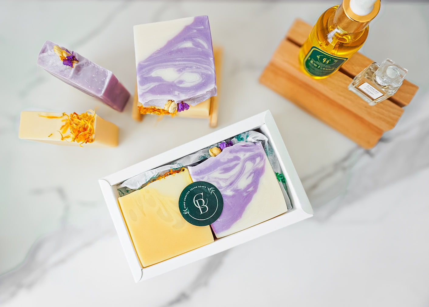 handcrafted soap bars (yellow or purple) in a white box and a oil and a mini perfume on a wooden stand