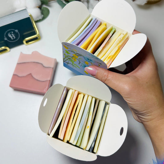 Hand holding a small open box with colorful soap end pieces and another box next to it. Full size regular soap as comparison.