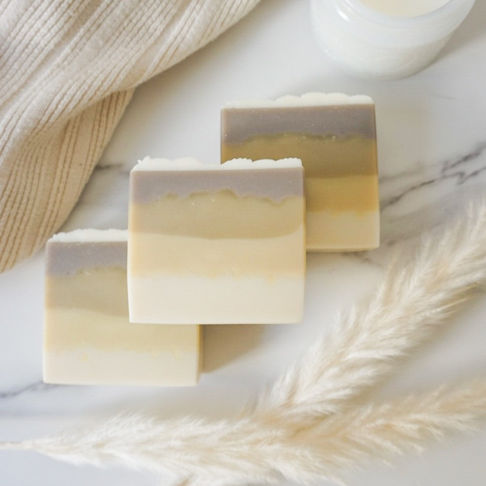 Three layered soap bars on a marble surface with a textured fabric and dried grasses.