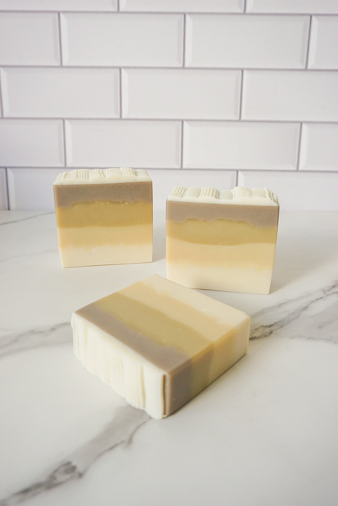 Three bars of soap with a gradient color on a marble surface with a tiled wall background.